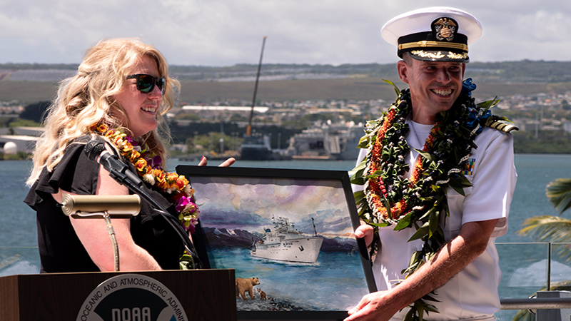 NOAA Ocean Exploration Operations Chief Kasey Cantwell presents outgoing commanding officer of NOAA Ship Okeanos Explorer, Captain Colin Little, with a painting of the ship that commemorates exploration done in Alaska in 2023 under CAPT Little’s command.